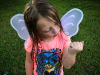 A photograph of a young girl with wet hair in a pink t-shirt watching a bug crawl on her closed hand. She is wearing a pair of translucent wings.