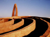 A photograph of an outdoor installation. This detail shows one edge of a series of low, concentric walls, the interiors in shadow, with a tall arc standing in the background