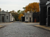 A photograph of a small-stone paved road, lined by ornate mausoleums, inside a tidy cemetary