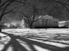 A black and white photo of the White House, seen with long tree shadows lain ominously across the snowy ground