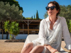 A photograph of a woman wearing sunglasses and a loose-fitting knee-length white garment sits on a lawnchair by a pool