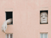 A photograph of a two windows on a two story apartment building. A face (oversized) fills one and a giant hand emerges from the other.