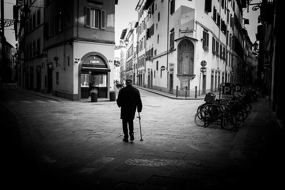 A black and white photo of a man using a cane staring at the buildings in an otherwise empty city