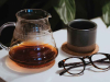 A photograph of a clear tea pot, a tea mug, and a pair of glasses on a table