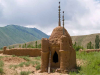 A tomb, built from clay, with rounded shapes suggestive of a mosque, sits in a sparsely covered grass field outside the walls of a city