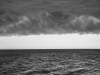 A black and white photo of the sea, dark clouds looming overhead, with the horizon forming the only light between them