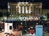 Looking from behind an outdoor stage towards a large crowd assembled to watch a performance with a large building in the background