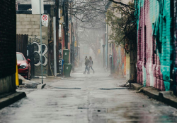 A photograph looking down a city alley. A couple holding hands strides away from the camera in the distance.