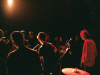 Three musicians stand on a lit stage in a darkened room with the audience standing just inches away