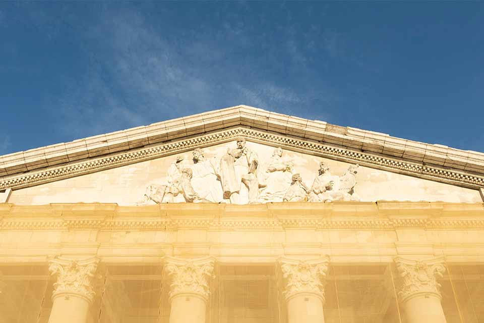 A photograph of a detail near the roofline of an aged marble building. The area has a relief sculpted into including several historical and mythological figures, including Joseph Conrad