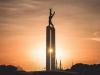 A photograph of a statue of a humanoid figure high on a tower. The tower is in the halo of a sunrise or sunset