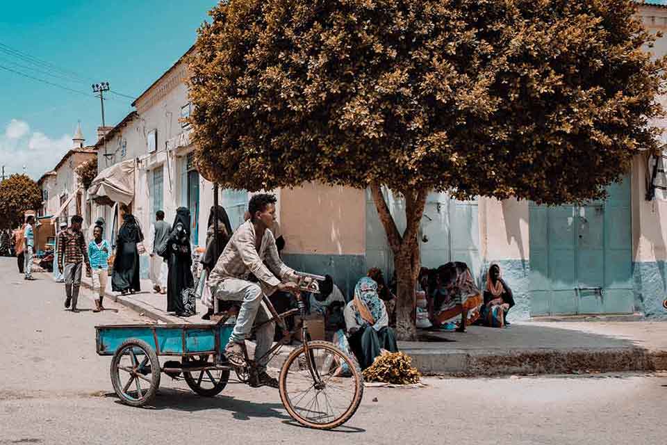 A photograph of a man riding a bicycle, pulling a small blue wagon, down a quiet street with people sitting and standing on the sidewalk. A tree offers shade to a few people sitting beneath it