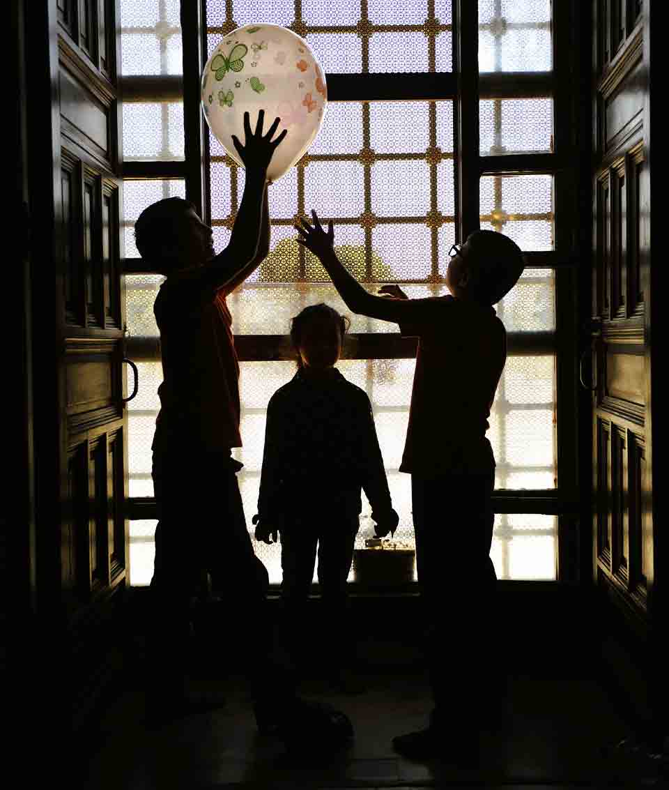 A photograph taken from inside a darkened building. Three children play with a balloon suspended in mid-air. The figures are draped in shadow, backlit by a window behind them.