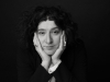 A black and white photograph of a woman, her head resting in her hands, looking soberly at the viewer