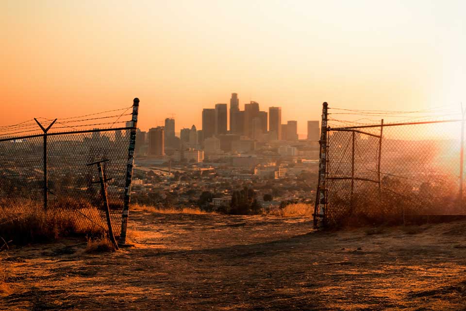 A photograph of a city from a distance, as seen through a ruined chain link fence. An orange haze sits over the city.