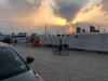 A young boy pauses on his bicycle on a gravel road with the sun either rising or setting behind a makeshift wall